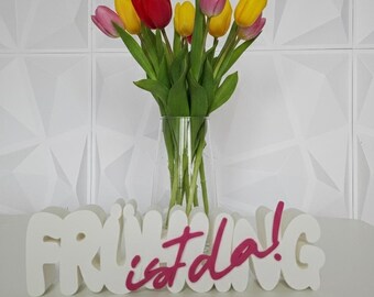 May include: A clear glass vase holds a bouquet of colorful tulips in shades of red, yellow, and pink. The vase sits in front of a white, 3D-lettered sign that reads "FRÜHLING ist da!" in a playful font, with the words "ist da!" in pink.