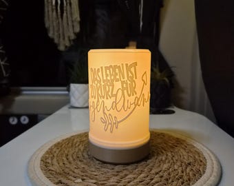 May include: A cylindrical, illuminated lamp with a warm glow, featuring the German text "DAS LEBEN IST ZU KURZ FÜR irgendwann" etched into its surface. The lamp has a solid, neutral-colored base and a smooth, translucent body.