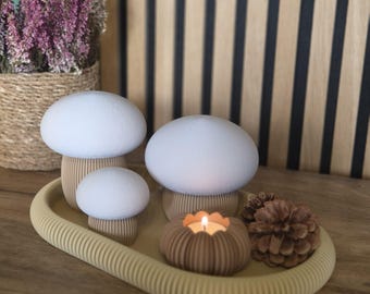 May include: A set of three white mushroom-shaped decorative objects with ribbed tan stems, arranged on an oval tan tray alongside a small ribbed candle holder with a lit tea light and two brown pinecones, creating a cohesive display.