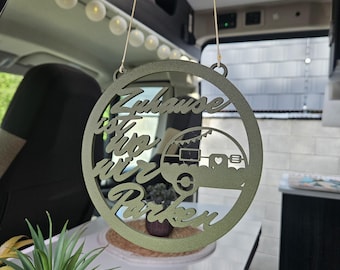 May include: A circular olive green hanging sign featuring a cutout design of a camper trailer and the German text "Zuhause ist wo wir Parken". The sign is suspended by two thin strings and displays a matte finish with a decorative bunting detail.