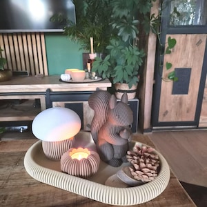 Könnte beinhalten: Ein ovales beiges Tablett mit einer weißen pilzförmigen Lampe, einer kleinen braunen Eichhörnchenfigur mit Eichel, einem runden gerippten Kerzenhalter mit brennendem Teelicht und einem natürlichen Kiefernzapfen, arrangiert auf einer Holzoberfläche.