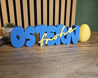 Puede incluir: Decoración de Pascua azul y amarilla. La palabra "OSTERN" es azul, con "frohe" en amarillo, y un huevo amarillo a la derecha. La decoración está sobre una superficie de madera.