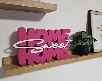 May include: A hot pink "HOME sweet HOME" sign with white cursive text, displayed on a wooden shelf. A small potted plant and a framed botanical print are also on the shelf. A candle in a glass jar sits on a shelf above.