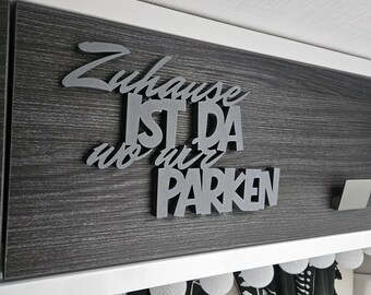 May include: A gray wooden sign with the German phrase "Zuhause ist da wo wir parken" (Home is where we park) mounted on a dark gray wood-grain surface. The sign is above a white shelf with decorative lights and black and white curtains.