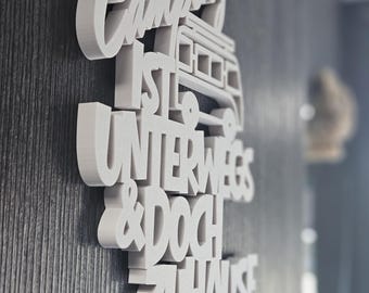 Puede incluir: Un adorno de pared 3D blanco con la frase alemana "Camping ist unterwegs & doch zuhause" (Acampando está de viaje y sin embargo en casa). El diseño incluye una ilustración de una furgoneta camper. Montado en una pared gris texturizada.