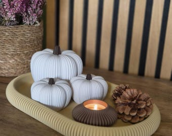 May include: An oval-shaped yellow tray with a ribbed edge holds two white pumpkins with dark brown stems, a small dark brown candle holder with a lit tea light, and two brown pinecones. The tray is set on a wooden surface.