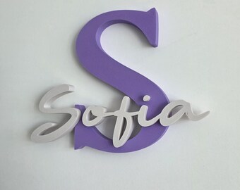 May include: A lavender-colored wooden letter "S" with the name "Sofia" in white script. The name is layered over the letter, creating a 3D effect. The background is plain white.
