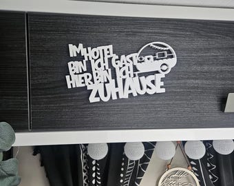 May include: White wooden sign with the German text "IM HOTEL BIN ICH GAST HIER BIN ICH ZUHAUSE" and a camper silhouette. The sign is mounted on a dark wood-grain surface, with black and white curtains and decorative lights below.
