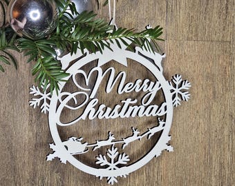 May include: White wooden Christmas ornament with the words "Merry Christmas" in a circular design. The ornament features snowflakes, reindeer pulling a sleigh, and a heart. Greenery and silver ornaments are visible in the background.