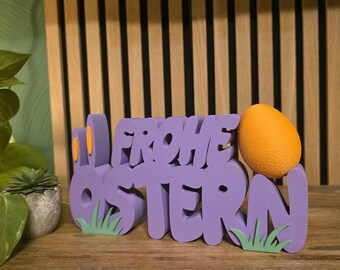 Puede incluir: Un letrero decorativo morado con el texto alemán "FROHE OSTERN" y un huevo naranja. El letrero tiene detalles de hierba verde y detalles naranjas. El letrero está sobre una superficie de madera.