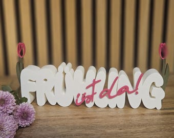 May include: A white wooden sign with the word "FRÜHLING ist da!" in a playful font, with pink accents. The sign is decorated with pink tulip cutouts and rests on a wooden surface. A bouquet of purple flowers is in the foreground.