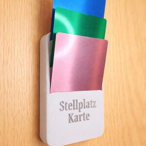 Può includere: Un porta carte bianco con tre carte colorate: blu, verde e rosa. Il supporto è montato su una superficie in legno e presenta la scritta "Stellplatz Karte".