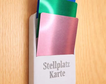 May include: A white card holder with three colorful cards: blue, green, and pink. The holder is mounted on a wooden surface and has the text "Stellplatz Karte" printed on it.