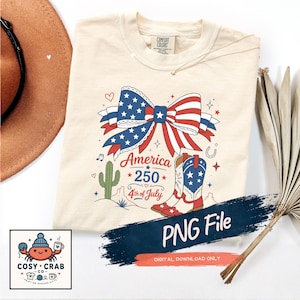 May include: A cream-coloured t-shirt featuring a graphic of a large bow with red, white, and blue stars and stripes, a cactus, and a cowboy boot. The text reads America 250 4th of July. A dark blue brushstroke overlay says PNG File.