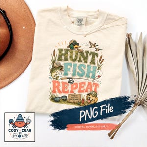 May include: A cream-colored t-shirt featuring a graphic with the text HUNT FISH REPEAT. The design includes a mallard duck in a cap, a bass, a golden retriever, a fishing rod, sunglasses, and a wooden sign that reads WAKE UP. HEAD OUT. MAKE MEMORIES.