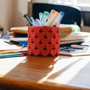 May include: A red heart-shaped pencil holder filled with colourful pens and markers. The holder is sitting on a wooden table with other writing utensils.
