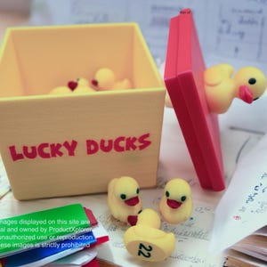 May include: A yellow box with the words "LUCKY DUCKS" printed on the front. The box is open and filled with yellow rubber duck toys. The box is sitting on a table with a stack of papers.