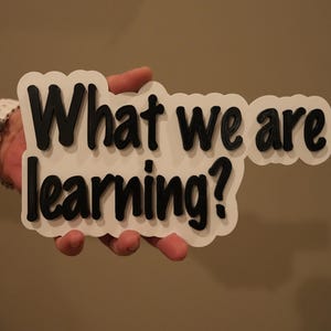 May include: White sign with black text that reads "What we are learning?"