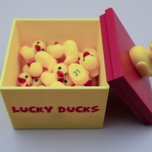 May include: A yellow box with the words "LUCKY DUCKS" printed on the front. The box is open and filled with yellow rubber duckies. The lid of the box is red and has a yellow rubber duckie attached to it.