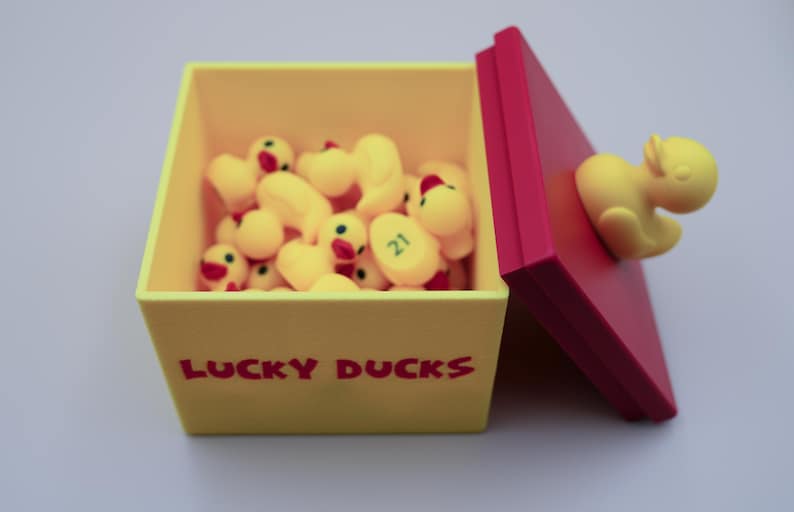 May include: A yellow box with the words "LUCKY DUCKS" printed on the front. The box is open and filled with yellow rubber duckies. The lid of the box is red and has a yellow rubber duckie attached to it.