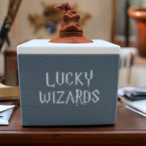 Pu&ograve; includere: Una scatola grigia con le parole "Lucky Wizards" stampate sul davanti. La scatola ha una statuetta di cappello di smistamento marrone in cima.