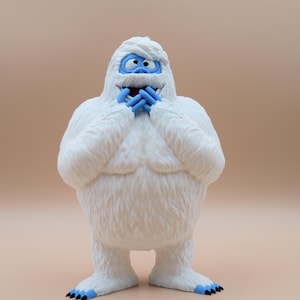 May include: A white, furry snowman figurine with a blue face and hands, smiling with its mouth open and hands covering its mouth. The figurine is standing on a light brown surface.