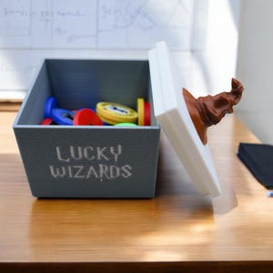 Pu&ograve; includere: Una scatola grigia con le parole "Lucky Wizards" stampate sul davanti. La scatola &egrave; piena di gettoni di plastica colorati e ha un coperchio bianco con un cappello da mago marrone in cima.