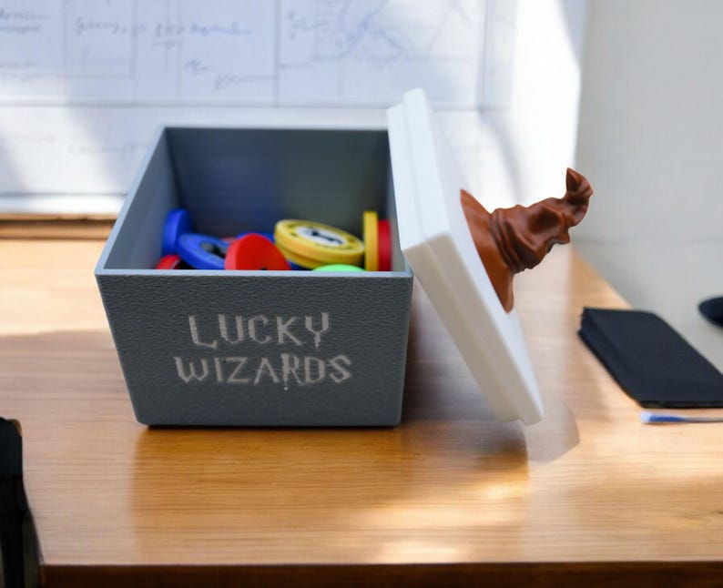 Pu&ograve; includere: Una scatola grigia con le parole "Lucky Wizards" stampate sul davanti. La scatola &egrave; piena di gettoni di plastica colorati e ha un coperchio bianco con un cappello da mago marrone in cima.