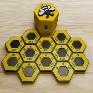 May include: A yellow multiplication game with a bee design. The game includes a yellow die with a bee and the letter "X", and yellow hexagonal tiles with multiplication problems like "3x6" and answers. The game is on a wooden surface.