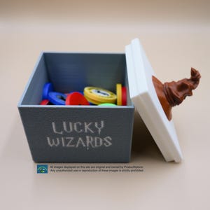 Pu&ograve; includere: Una scatola grigia con le parole "LUCKY WIZARDS" stampate sul davanti. La scatola &egrave; aperta e contiene una variet&agrave; di gettoni colorati. Il coperchio della scatola &egrave; bianco e ha un cappello da mago marrone in cima.