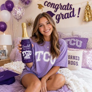 May include: A decorated champagne bottle with the text "Scarlett Class of 2026 TCU" is held up. The bottle is purple with gold accents. The person is wearing a purple TCU t-shirt. Balloons and a graduation banner are in the background.