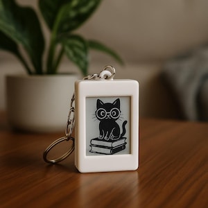 Könnte beinhalten: Ein weißer, rechteckiger Schlüsselanhänger mit einer silbernen Kette und einem Ring. Der Schlüsselanhänger zeigt eine schwarz-weiße Illustration einer Katze mit Brille, die auf Büchern sitzt. Der Hintergrund ist hellgrau.