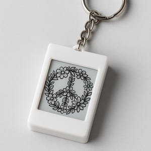 Peut inclure: Porte-cl&eacute;s rectangulaire blanc avec une cha&icirc;ne et un anneau argent&eacute;s. Le porte-cl&eacute;s pr&eacute;sente un insert gris avec un motif de signe de paix floral noir. Le signe de paix est entour&eacute; d'une couronne de fleurs et de feuilles.