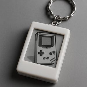 Peut inclure: Porte-cl&eacute;s blanc avec une image en niveaux de gris d'une Game Boy. Le pendentif rectangulaire a des coins arrondis et un anneau en m&eacute;tal argent&eacute;. Le graphisme de la Game Boy comprend le mot "GAMEBOY" et des boutons de commande.