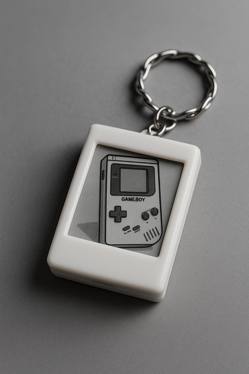 Peut inclure: Porte-cl&eacute;s blanc avec une image en niveaux de gris d'une Game Boy. Le pendentif rectangulaire a des coins arrondis et un anneau en m&eacute;tal argent&eacute;. Le graphisme de la Game Boy comprend le mot "GAMEBOY" et des boutons de commande.