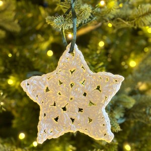 May include: A white crocheted star ornament hanging on a Christmas tree with green branches and white fairy lights.