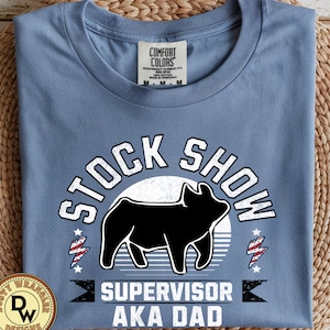Può includere: Una maglietta blu Comfort Colors con la scritta "STOCK SHOW SUPERVISOR AKA DAD". Il design presenta una silhouette di maiale nero, un cerchio bianco e dettagli rossi e blu. La maglietta è piegata su una superficie tessuta.