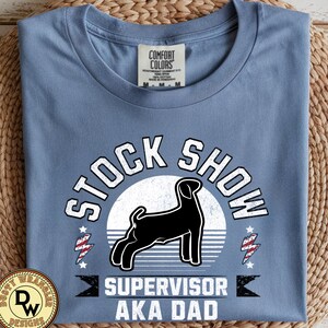 Può includere: T-shirt blu Comfort Colors con la scritta "STOCK SHOW SUPERVISOR AKA DAD". Il design presenta una silhouette nera di cane, un cerchio bianco e grafiche a fulmine. La maglietta è piegata.