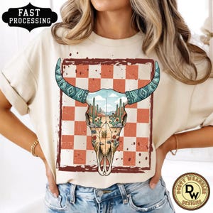 May include: Cream-colored t-shirt featuring a bull skull design with turquoise horns and a desert landscape inside. The skull is set against a red and white checkered background. The shirt also has the text "FAST PROCESSING".