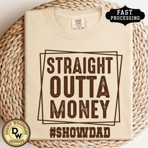 May include: A beige Comfort Colors t-shirt with the text "STRAIGHT OUTTA MONEY" in brown, framed by a brown rectangle. Below the text is the hashtag "#SHOWDAD". The shirt has a round neckline and short sleeves.