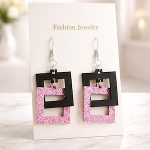 May include: A pair of dangling earrings with a black square frame over a pink glitter square. The earrings are displayed on a white card with the words "Fashion Jewelry" printed in gold. The earrings hang from silver hooks.