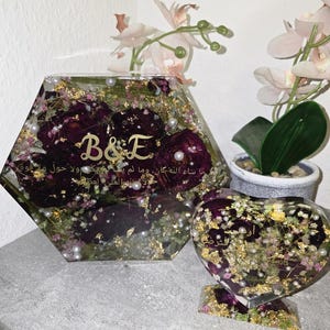 Op de afbeelding: Twee harskunstwerken, een zeshoekig en een hartvormig, gevuld met bordeauxrode bloemen, goudvlokken en parels. Het zeshoekige stuk heeft "B&E" en Arabische tekst. Het hartvormige stuk heeft ook Arabische tekst.