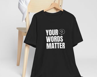May include: Black t-shirt with the white text "YOUR WORDS MATTER" and a heart symbol inside a speech bubble. The shirt is hanging on a wooden hanger. A white baseball cap rests on the back of a wooden chair.