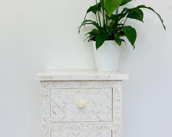 May include: White wooden nightstand with two drawers featuring intricate floral patterns and white knobs. The nightstand is sitting on a patterned rug.
