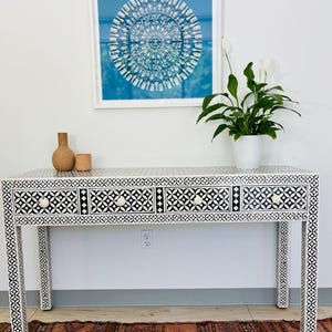 May include: A black and white bone inlay console table with four drawers and white knobs. The table has a geometric pattern and is sitting on a rug with a similar pattern. There is a framed print of a blue mandala above the table.