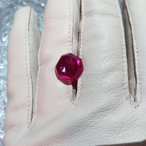 Può includere: Una pietra preziosa di colore rosso intenso con taglio sfaccettato, esposta su una superficie di pelle bianca.