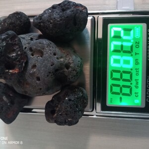May include: A digital scale with a green display showing a weight of -8.8.8.8. The scale is displaying units of ct, dwt, ozt, gn, T, OZ. The scale has a capacity of 1000G X 0.1G. A pile of black volcanic rocks are on the scale.