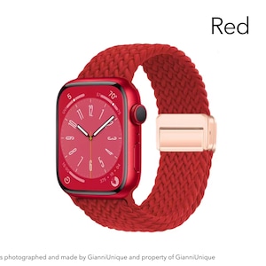 May include: A red smartwatch with a rectangular face and a matching red braided strap. The watch features a digital display with white hands and markers, a black side button, and a metallic rose gold-colored clasp on the woven band.