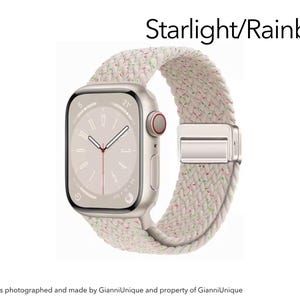 May include: A smartwatch with a rectangular starlight-colored case and a braided band featuring a starlight base with small, scattered rainbow-colored flecks. The watch face displays a minimalist analog clock design with the text Starlight/Rainbow.