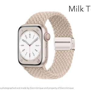 May include: A smartwatch with a rectangular gold-tone case and a beige braided fabric band featuring a metallic clasp. The watch face displays a white dial with black numerals and hands. The text Milk Tea is visible in the upper right corner.
